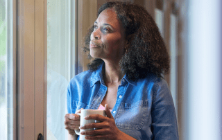 Black woman looking hopefully out the window while holding a coffee mug